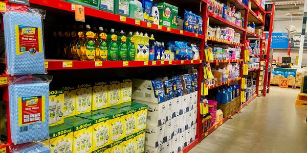 Store aisle stocked with household cleaning products, dishwashing liquids, wipes and detergents arranged on red shelving with price labels throughout.