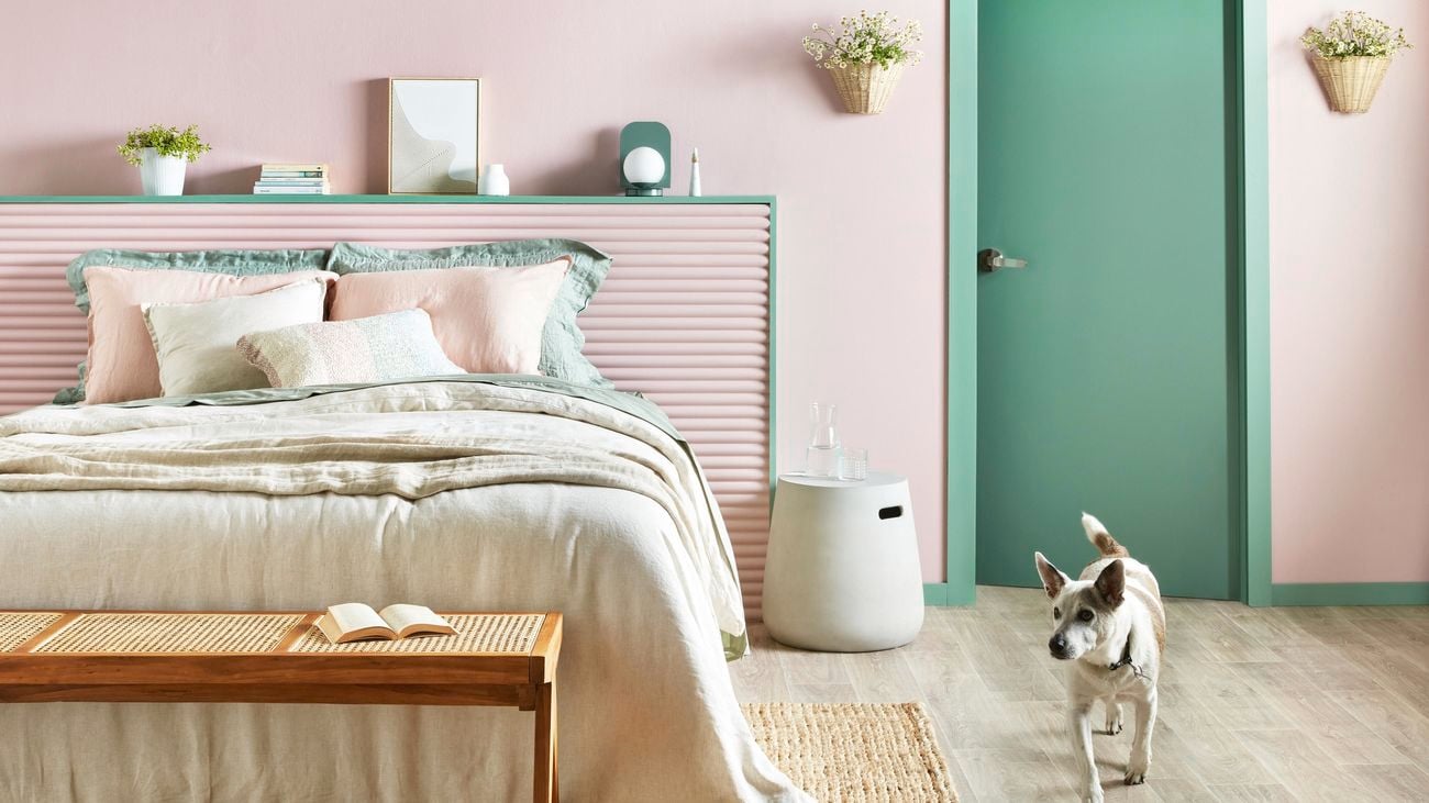 Bedroom with light pink walls and a fully made cream colour bed and wooden end-of-bed bench. There is a hanging wall vase on the pink wall
