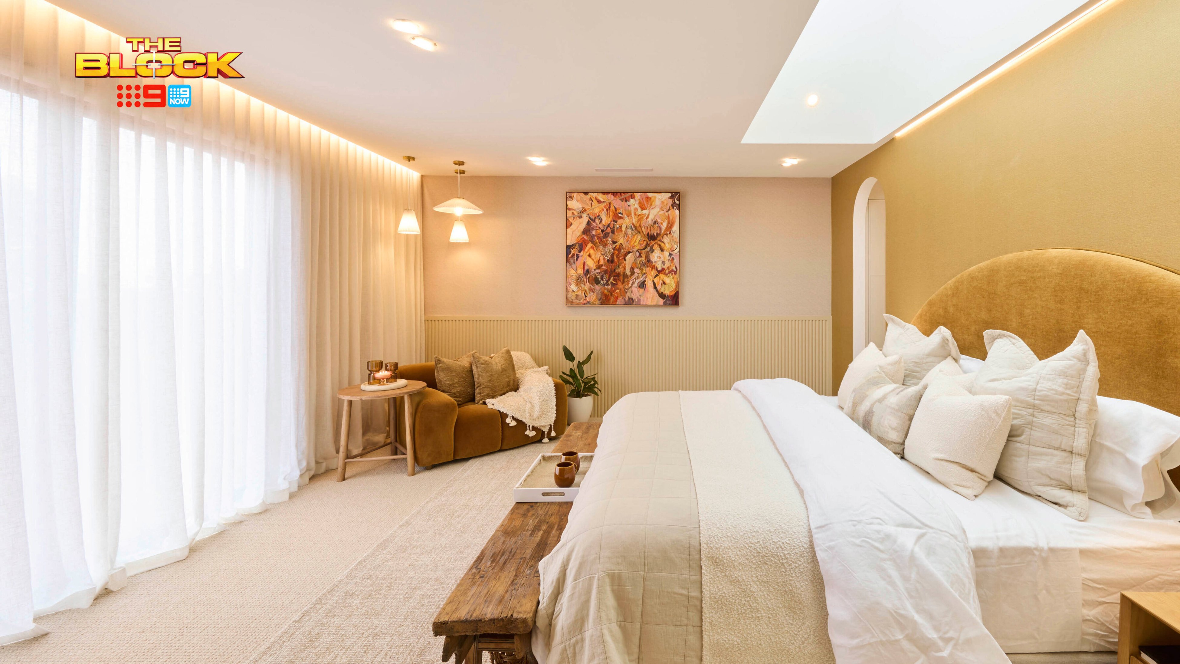A stylish bedroom featuring a large bed with multiple pillows and a beige color palette. A wooden bench sits at the foot of the bed. To the left, there’s a cozy seating area with a brown sofa, cushions, and two small tables. Large windows with sheer white curtains let in natural light. The walls are decorated with artwork and lighting fixtures. 