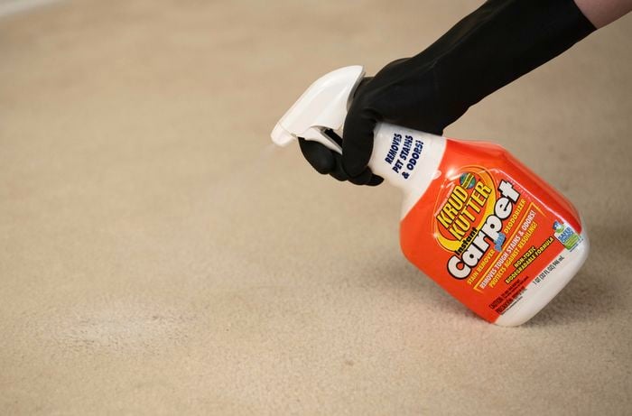 A person wearing black gloves is spraying a carpet stain remover plus deodoriser onto a stained beige carpet.