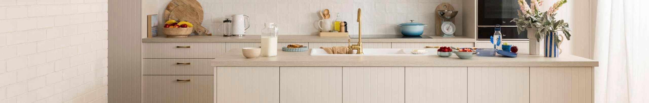 A beige modern kitchen, with a brown benchtop with crockery on it, an oven in it and a table and chairs in front of it.