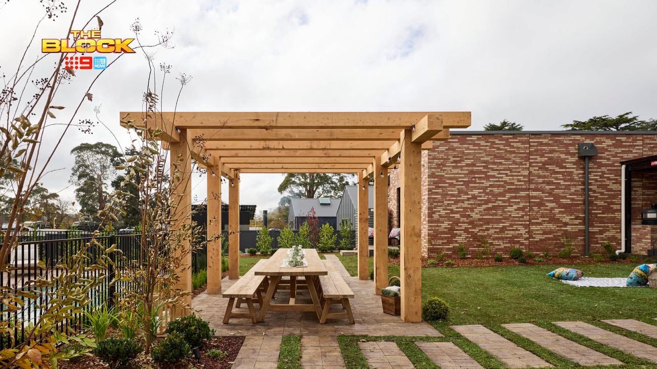 A backyard with paved steps amongst the grass, a pool, outdoor table with searing and a timber structure above it.