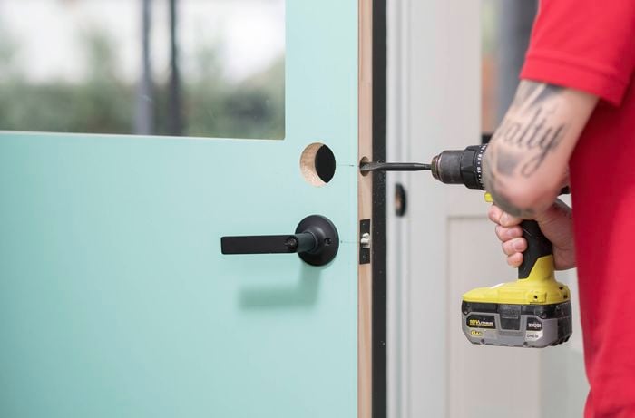 A person is drilling a hole into the edge to install a latch using a drill bit.
