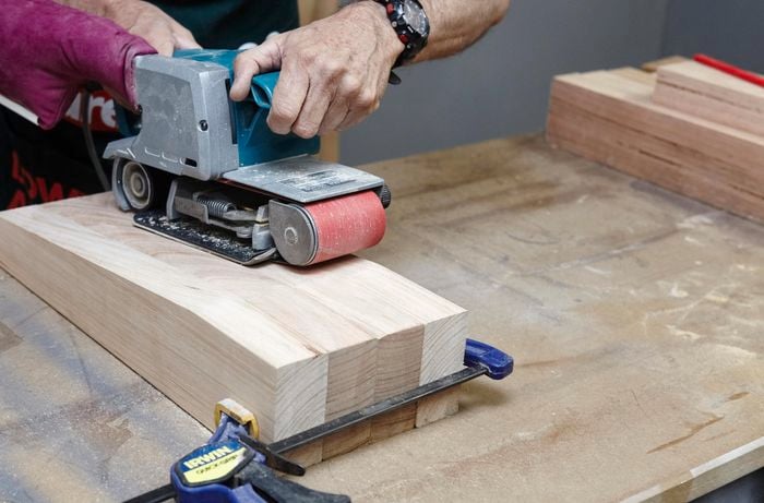 The four legs of a wooden chair being sanded down with a belt sander, held together with a clamp