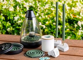 Close up of a timber outdoor table with greenery behind with natural mosquito repellent, mosquito coils plus a candle and plug-in diffuser on top