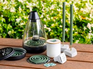 Close up of a timber outdoor table with greenery behind with natural mosquito repellent, mosquito coils plus a candle and plug-in diffuser on top