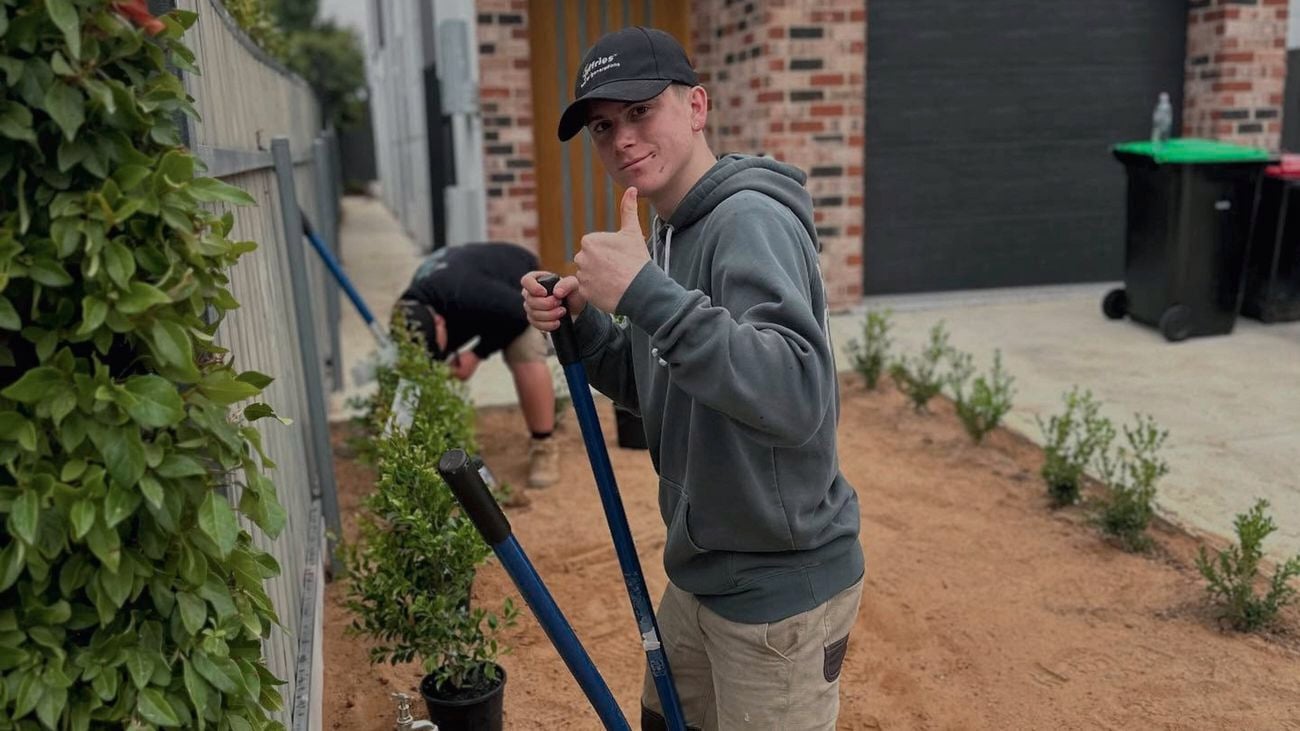 A person holding garden tools stands beside newly planted shrubs along a fence in a residential yard. Another person works in the soil nearby, with brick walls, a driveway, and trash bins visible in the background.