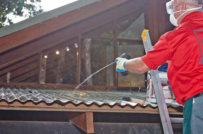 Person on a ladder wearing protective clothing spraying roof with a cleaning solution