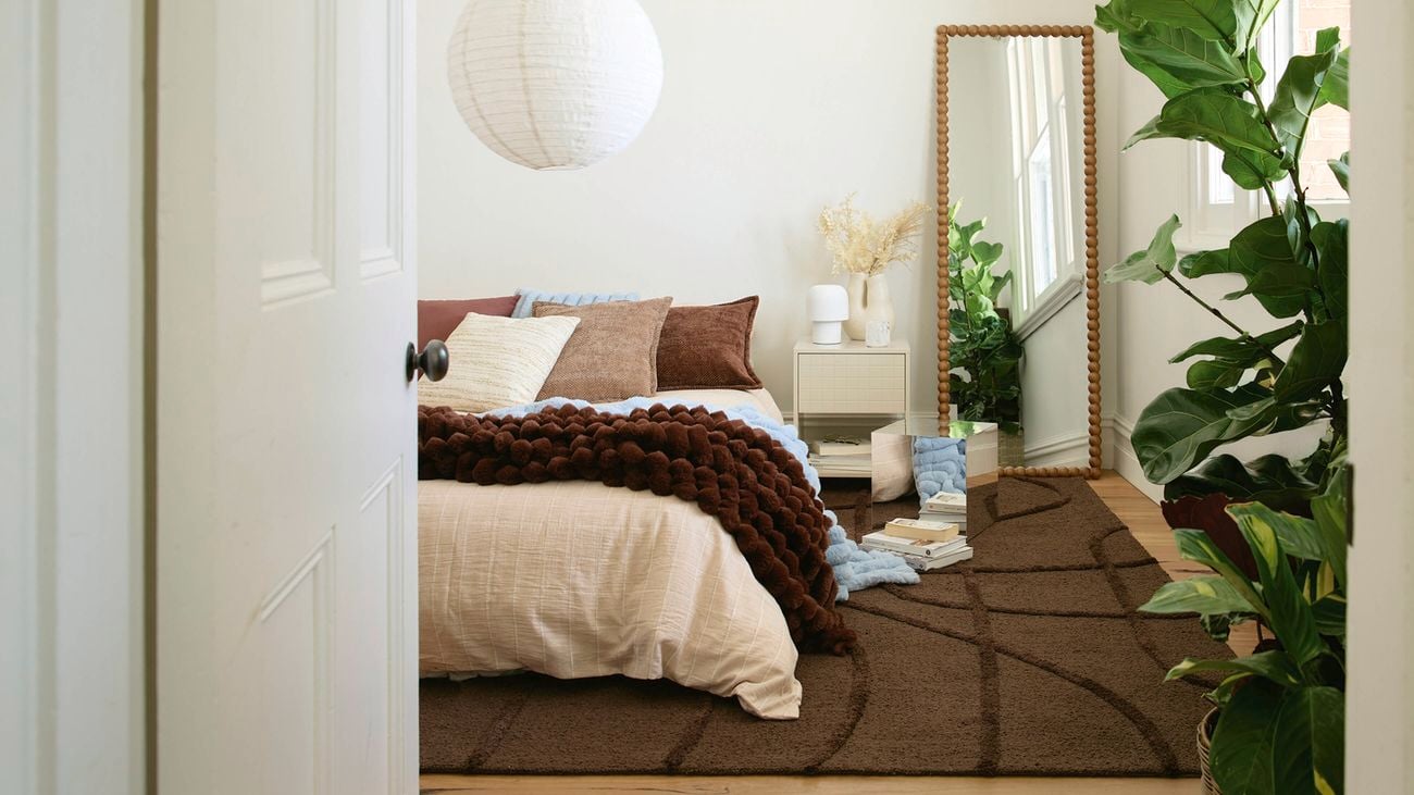 A bedroom with a bed, brown blanket, and pillows. A white paper lantern, bedside table with lamp and dried flowers, and a large beaded mirror reflecting a window and plants.]
