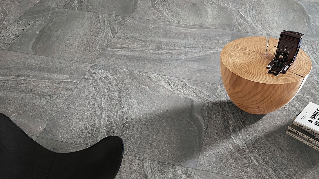 Grey tiled flooring, a wooden round coffee table with a camera on top, and a stack of books on the floor. Part of a black chair is visible in the foreground.