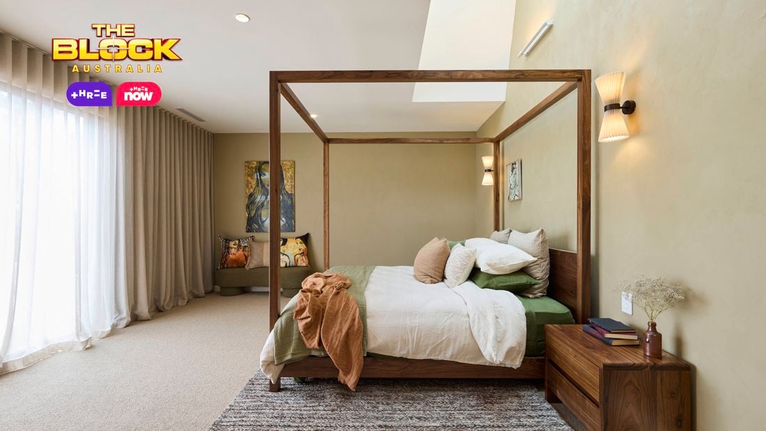 A stylish bedroom featuring a central four-poster bed with white and green bedding, multiple pillows, and a brown throw blanket. A wooden nightstand with books and decor sits to the right, while a cushioned bench with decorative pillows lines the left wall. The room includes a textured rug, light beige walls, wall-mounted lights, and artwork above the bench and bed. Sheer curtains cover large windows, allowing natural light in. 