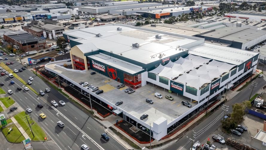 Aerial view of a Bunnings store surrounded by a large parking lot with rows of cars. The store is a large warehouse-style building with a prominent sign displaying the Bunnings logo.