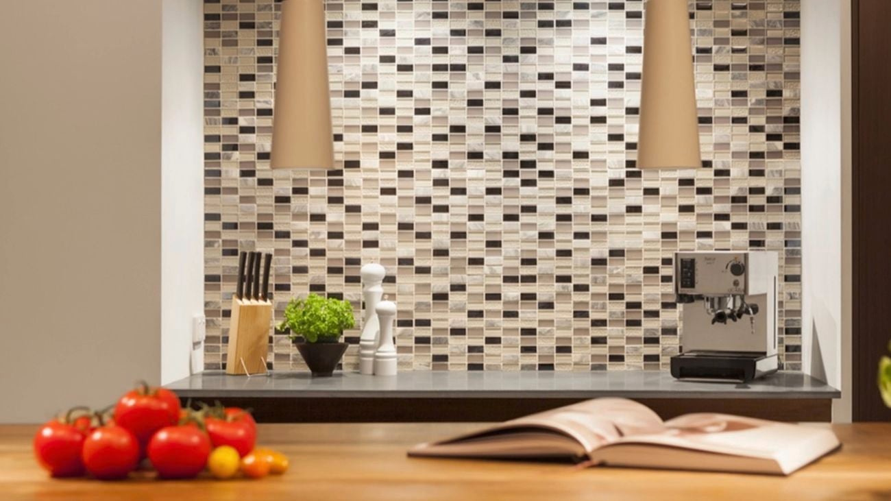 Kitchen benchtop with a mosaic tile splashback, pendant lights overhead, and styled accessories including tomatoes, a cookbook, knives, plants and a coffee machine.