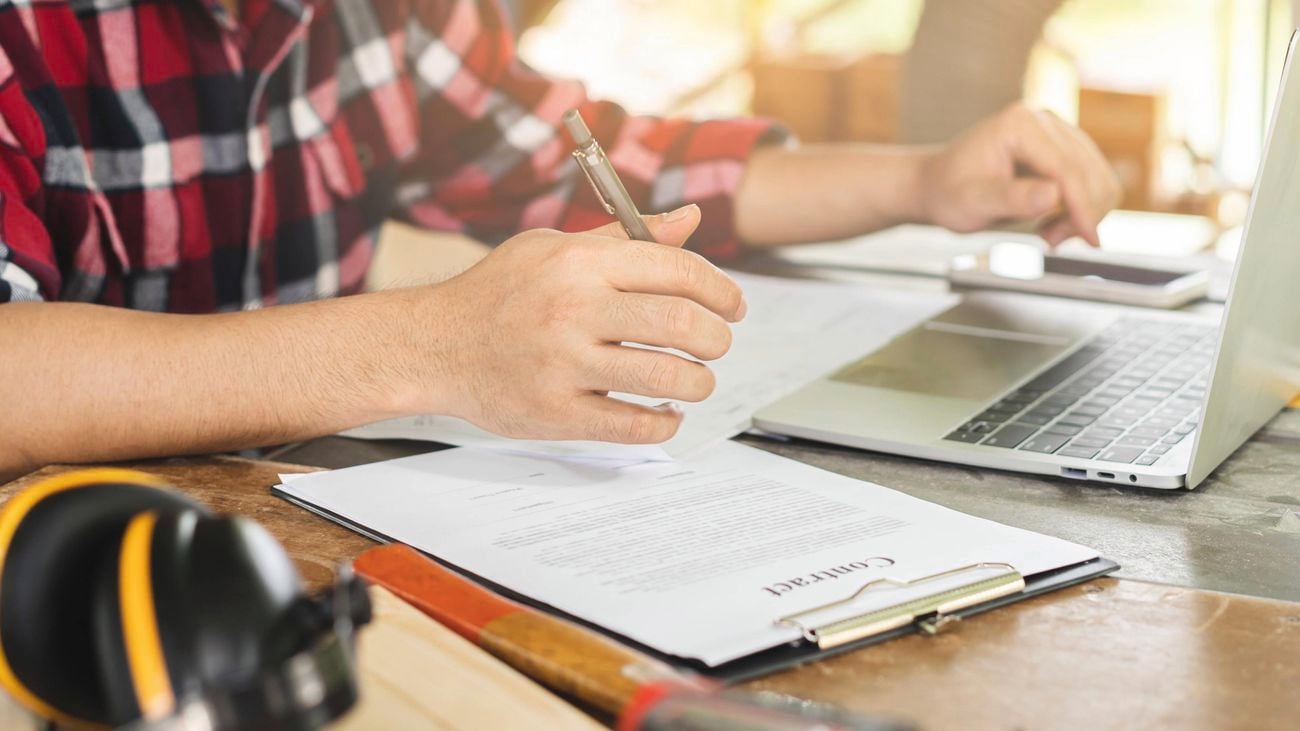 A person's hands are seen working on a contract and a laptop, with earmuffs and a hammer on the desk.