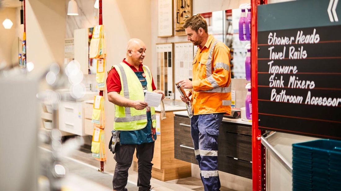 A tradie and a Bunnings Team Member in high-visibility clothing have a conversation in the plumbing section instore.