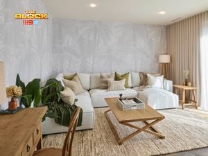 A modern living room with a large white sectional sofa decorated with neutral and green pillows. A wooden coffee table with a decorative tray sits in front, and a side table with a lamp and vase is placed to the right. Light-colored curtains cover large windows, and palm tree wallpaper adds texture to the walls. A desk with drawers and a chair is positioned on the left, near potted plants. 