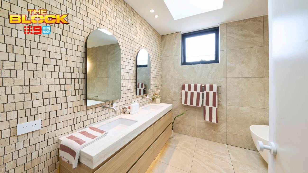 A modern bathroom, with a bath in it, a window, two mirrors on the wall, two basins with drawers underneath them and two towels hanging on a towel rail.