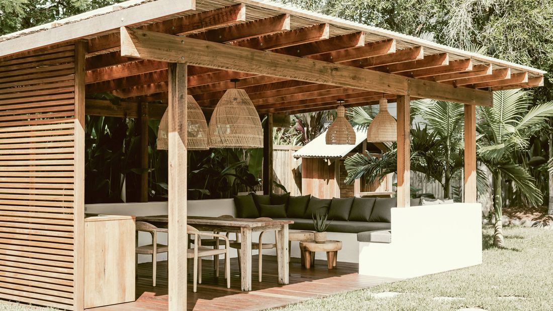  Outdoor living area with a wooden pergola, L-shaped sofa, dining table and lush tropical greenery.