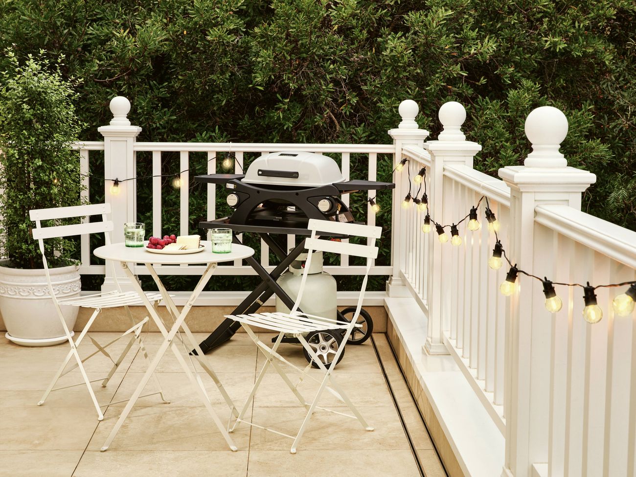 Outdoor entertaining area featuring a compact gas barbecue on a tiled deck, with a white bistro table set with drinks and snacks, white folding chairs, and warm string lights hanging along a white balustrade against a backdrop of dense greenery.