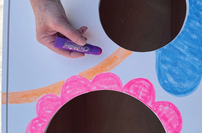 A hand holding a purple crayon near a white-painted timber surface with colourful drawings and two circular cut-outs.