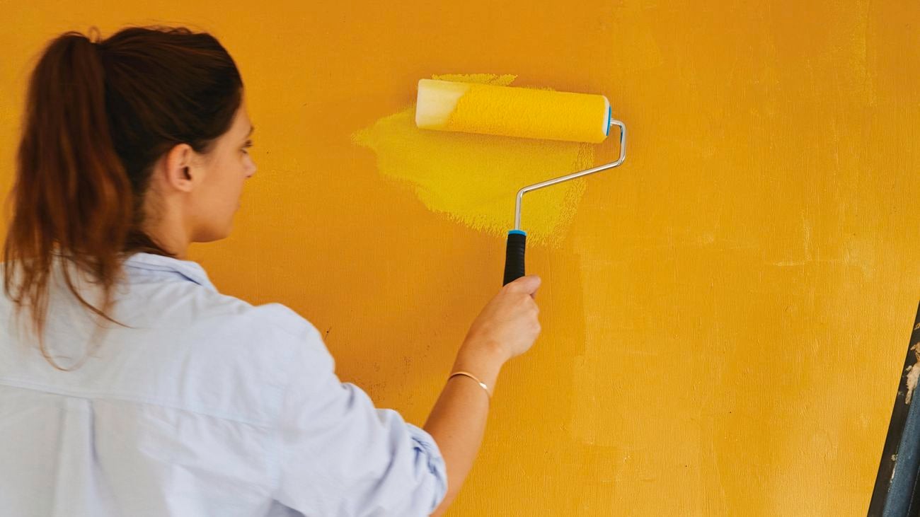 Woman with a brunette ponytail, a blue blouse and blue jeans using a paint roller to paint a wall mustard yellow.