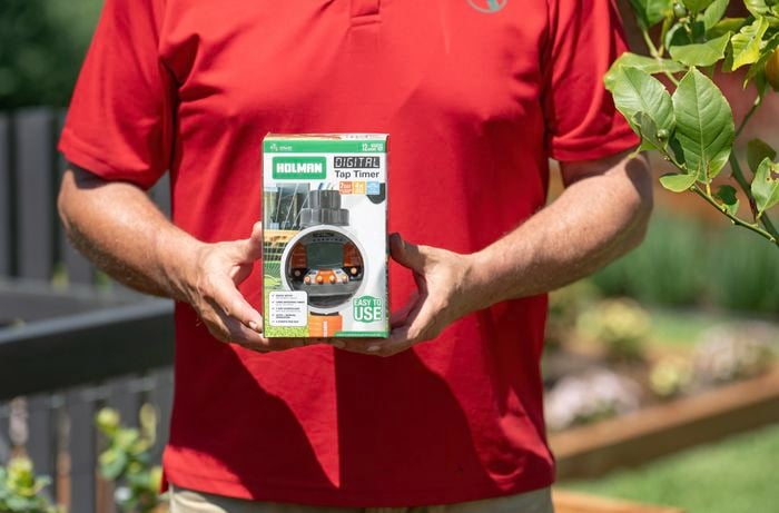 A Bunnings Team Member is holding a Holman digital tap timer in a garden, allowing for scheduled watering.