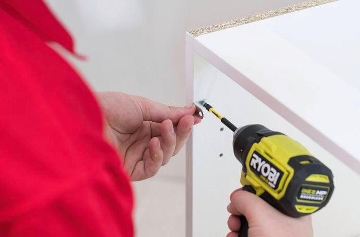 A person in a red t-shirt uses a drill to attach an L bracket to the top of the blind panel, with three brackets fixed at the bottom and two on the side. 