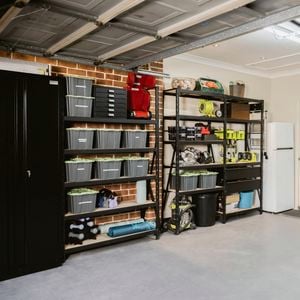 Organized garage with shelves holding plastic bins and tools, a black cabinet on the left, and a white refrigerator against the back wall. The open garage door reveals ceiling tracks and opener mechanism. 