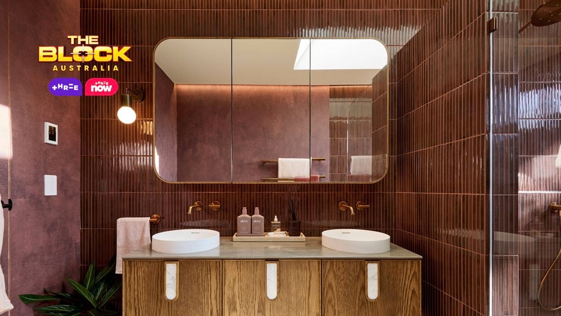 Modern bathroom with double round sinks on a wooden vanity, large mirror, maroon tiled walls, and partial view of a glass shower. 
