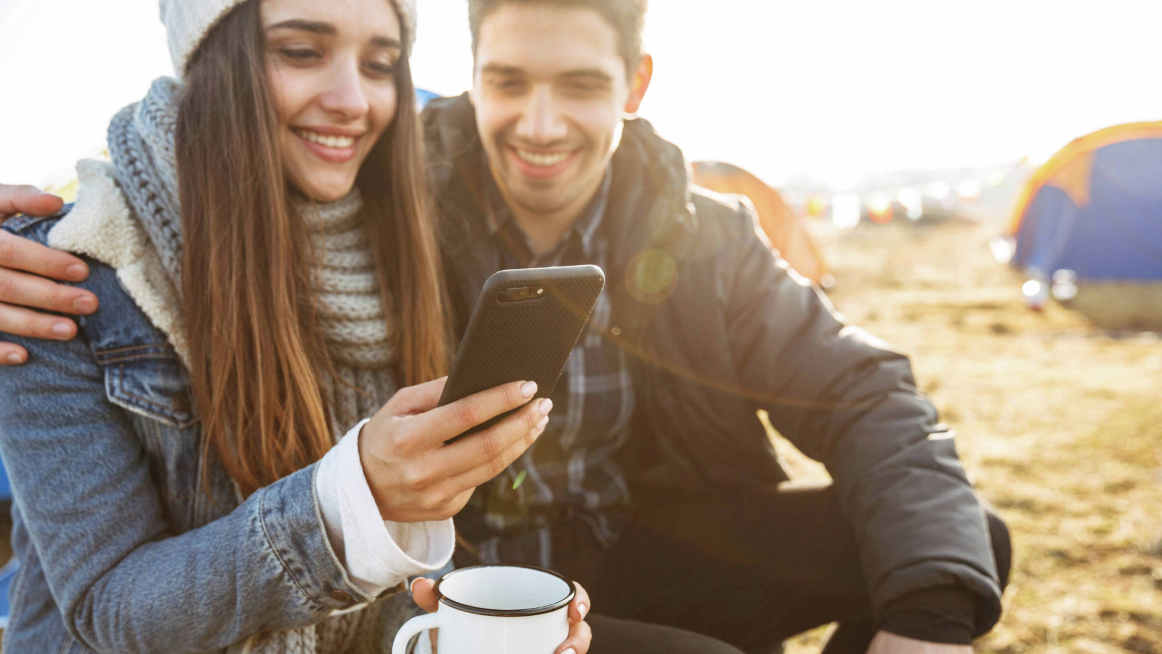 A woman is reading something on her smartphone, holding in front of her while holding a mug on the other hand accompanying with a man with his one hand over her shoulder on a camping background.