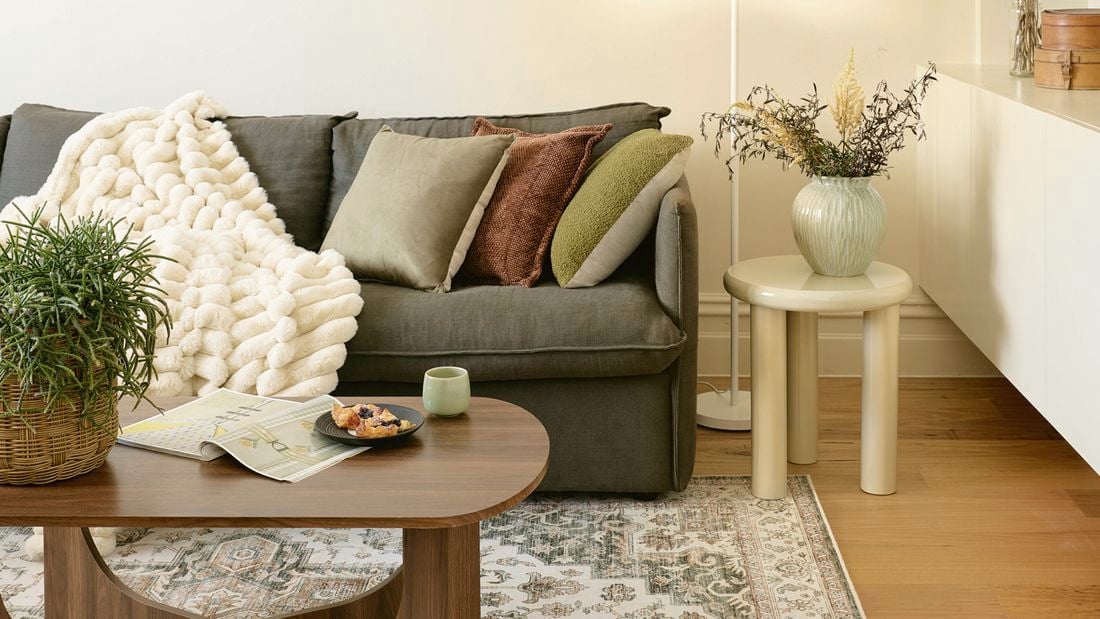 A cosy and warmly lit living room with a sofa with cushions and a throw, a wooden coffee table, shelving and a patterned rug