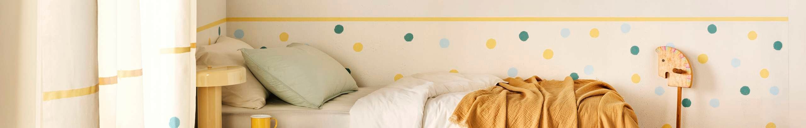 A children’s bedroom featuring a white bed with pastel bedding, a yellow throw, and a wall decorated with a yellow stripe and colourful polka dots. A hobby horse toy stands against the wall, and sunlight filters through curtains with matching yellow and blue accents.