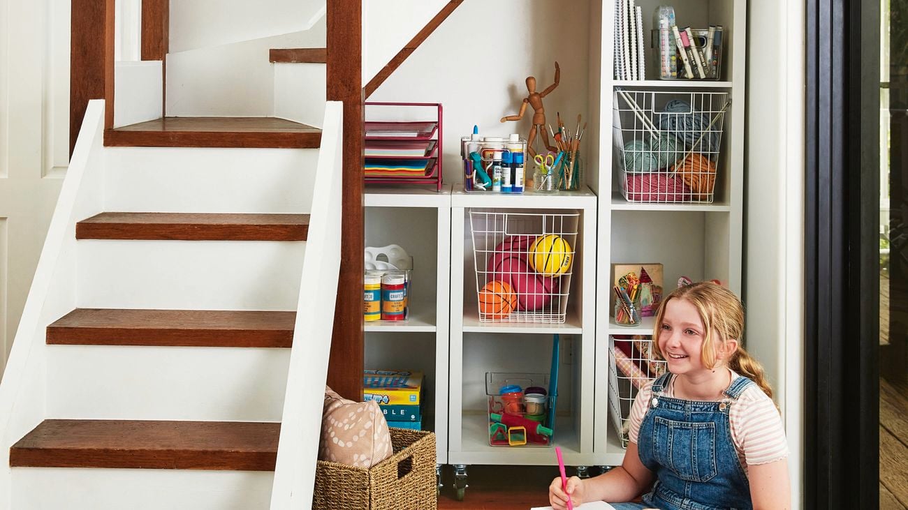 A nook under a staircase features white cube shelving filled with paper, paint supplies, scissors, notebooks and books. Wire baskets contain balls and yarn. A wicker basket is on the floor, and a young person in denim overalls and a striped shirt is seated on the floor, holding a pink pen and paper