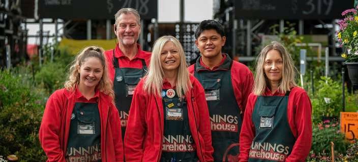 Group picture of five Bunnings team members dressed in Bunnings red uniform and green apron standing together in the outdoor garden section of a Bunnings store looking at the camera and smiling.