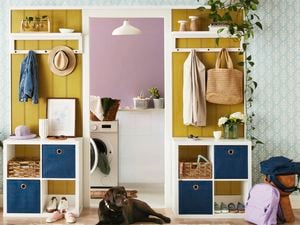 An entryway with a yellow wall and white cube shelving holding a straw hat, denim jacket, and woven baskets. A black Labrador dog lies on the floor, surrounded by shoes and a purple backpack. In the background is a laundry room with a washing machine and purple wall.