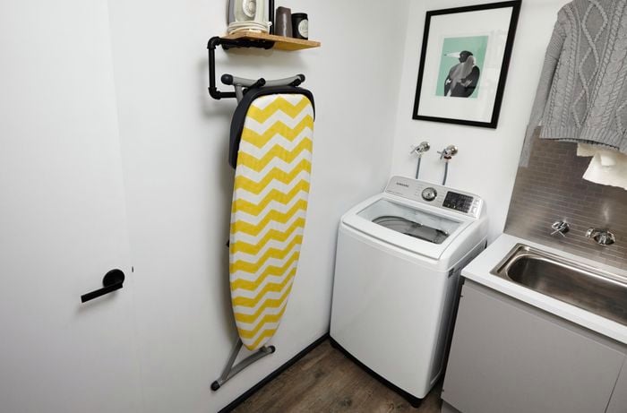 An ironing board storage rack mounted on a white laundry wall, with an ironing board hanging from it