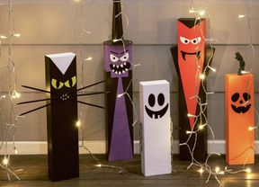 Halloween decorations made from painted rectangular blocks, including a cat, witch, ghost, vampire, pumpkin, and Frankenstein's monster, surrounded by string lights.