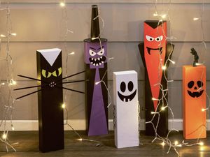Halloween decorations made from painted rectangular blocks, including a cat, witch, ghost, vampire, pumpkin, and Frankenstein's monster, surrounded by string lights.