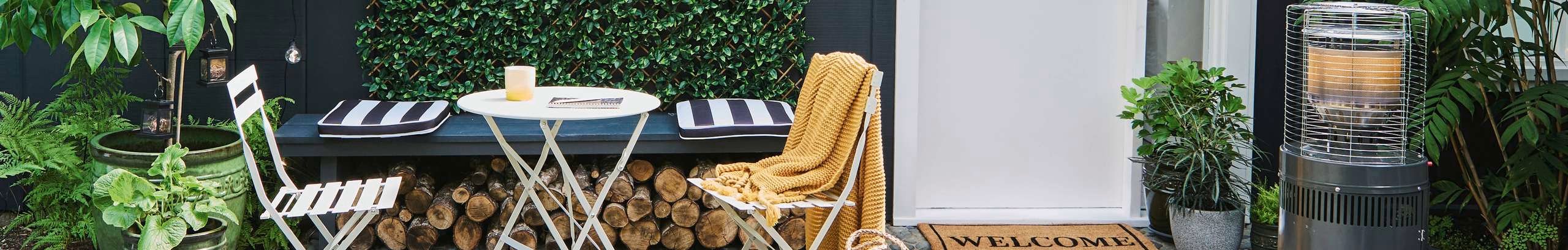 Outdoor patio area featuring a small white table with striped cushions on a bench, potted plants, a stack of firewood, and a yellow blanket draped over a chair. A welcome mat sits in front of a doorway, and a tall outdoor heater is positioned to the right.