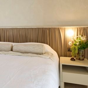 A furnished bedroom with a bed, side table with a vase on it and a bedhead behind it. And The Block logo in the image.