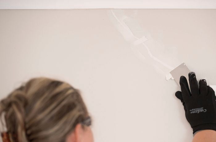 A person wearing black gloves is applying a second coat of filler with a putty knife to cover a repaired crack on a wall.