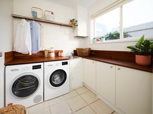 Laundry room with front-loading washer and dryer under a wooden benchtop, hanging shirts on a rail above, white cupboards, a sink in the corner, and a large window letting in natural light with indoor plants.