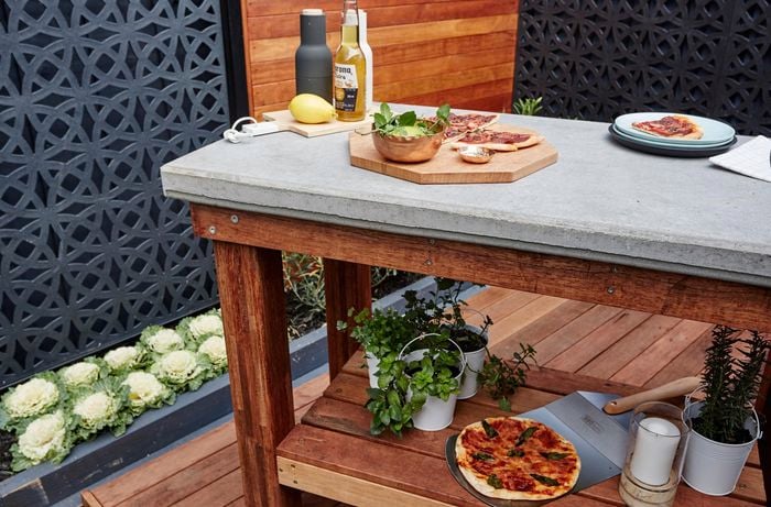 Outdoor table with food on tabletop and herbs on shelf beneath.