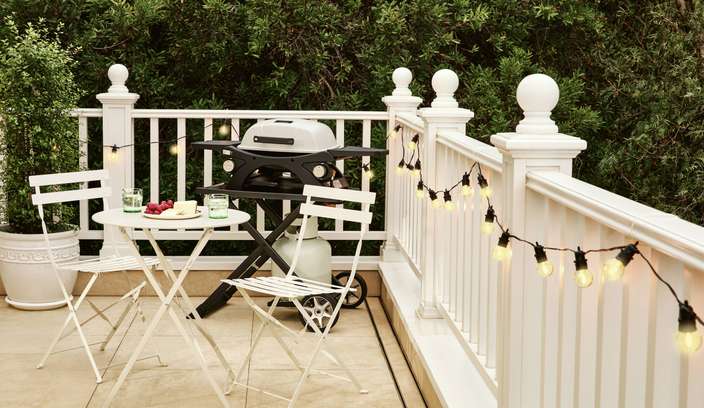 Deck with white balustrade, string lights, a gas barbecue, and a small outdoor dining set surrounded by greenery.