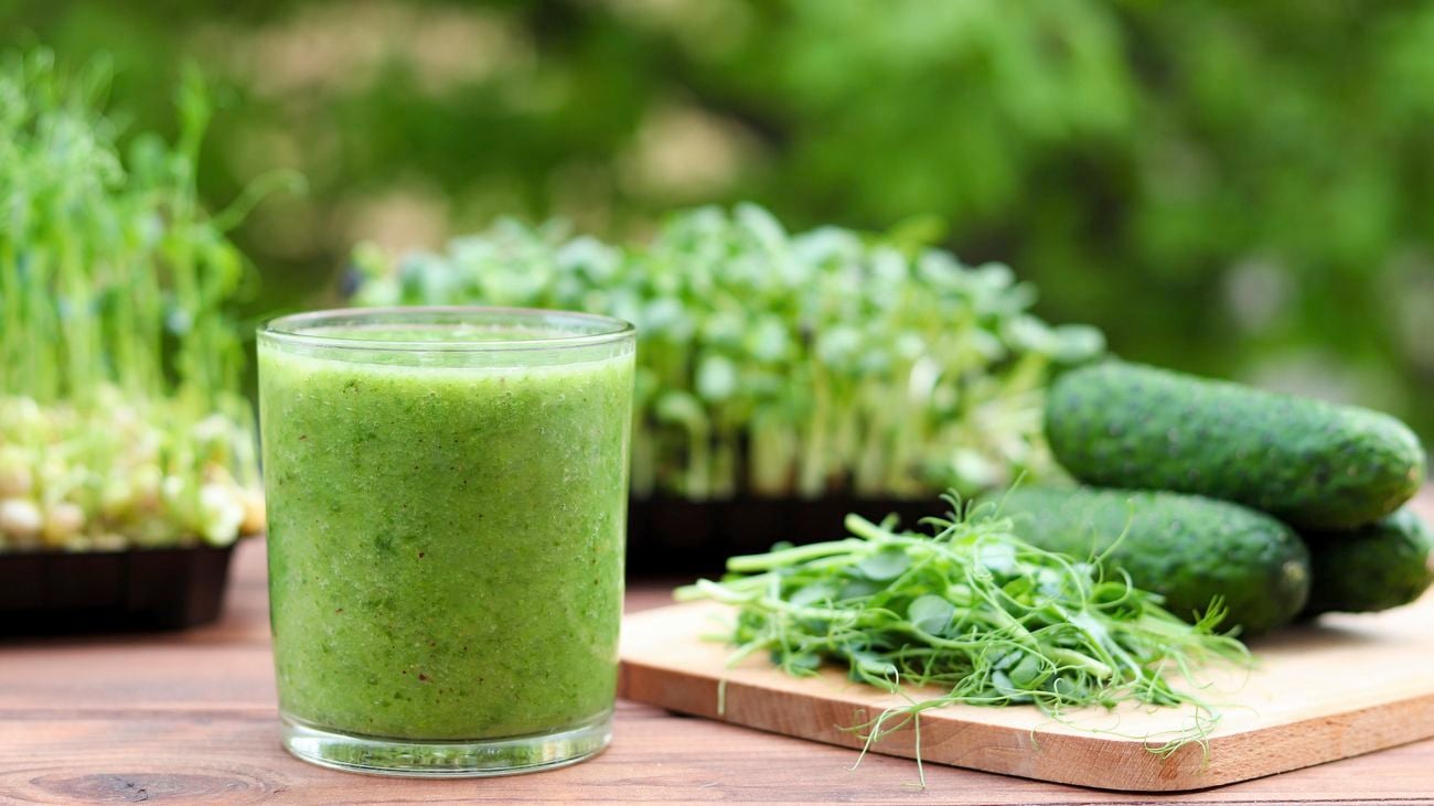 A green vegetable smoothie in a glass next to green vegetables on a wooden cutting board and microgreen plants in the background.