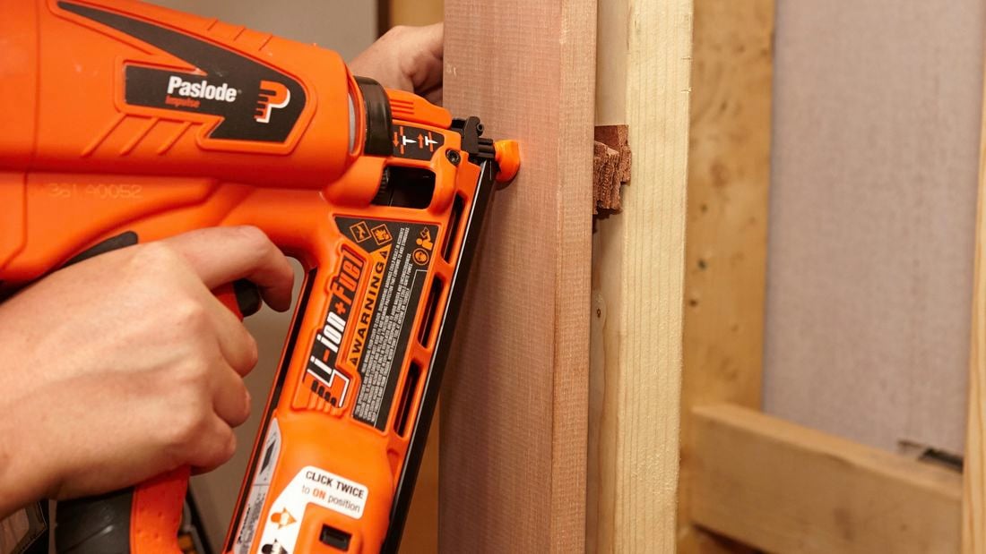 A person using a nail gun to attach a door frame to a wall stud with packers in between