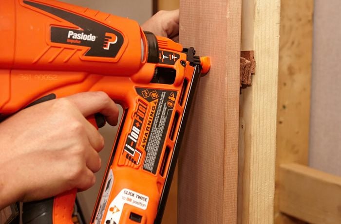 A person using a nail gun to attach a door frame to a wall stud with packers in between