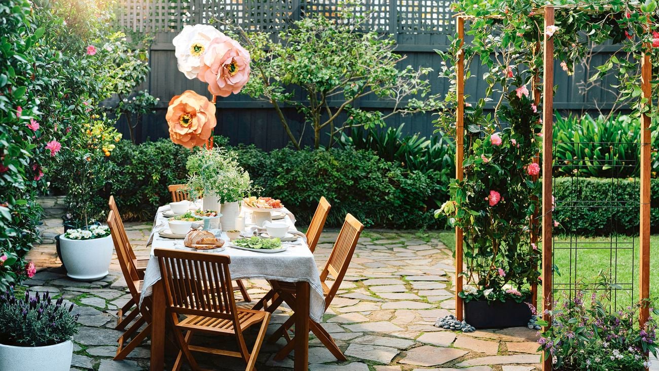 An outdoor dining table set in a garden courtyard beside a flower-covered garden arch and oversized paper flowers.