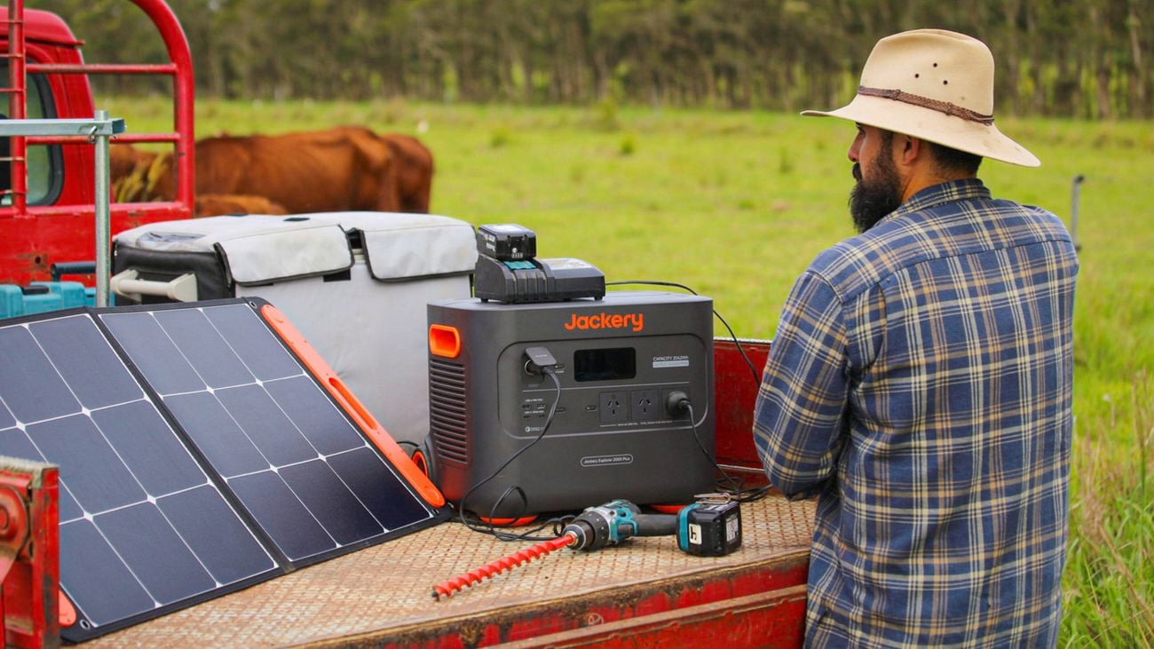 A man in a plaid shirt and hat stands next to a portable solar panel and Jackery power station on the tray of a red utility vehicle in a grassy field.