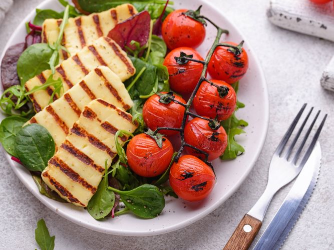 Grilled halloumi slices and charred cherry tomatoes served on a bed of mixed leafy greens in a white bowl.